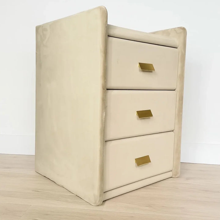 Beige or cream bedside table with three drawers and gold handles on light wood floor